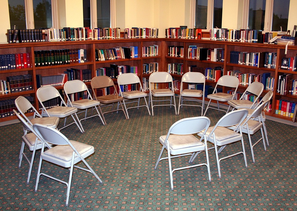 A circle of empty chairs on a carpet with a bookshelf behind them. 