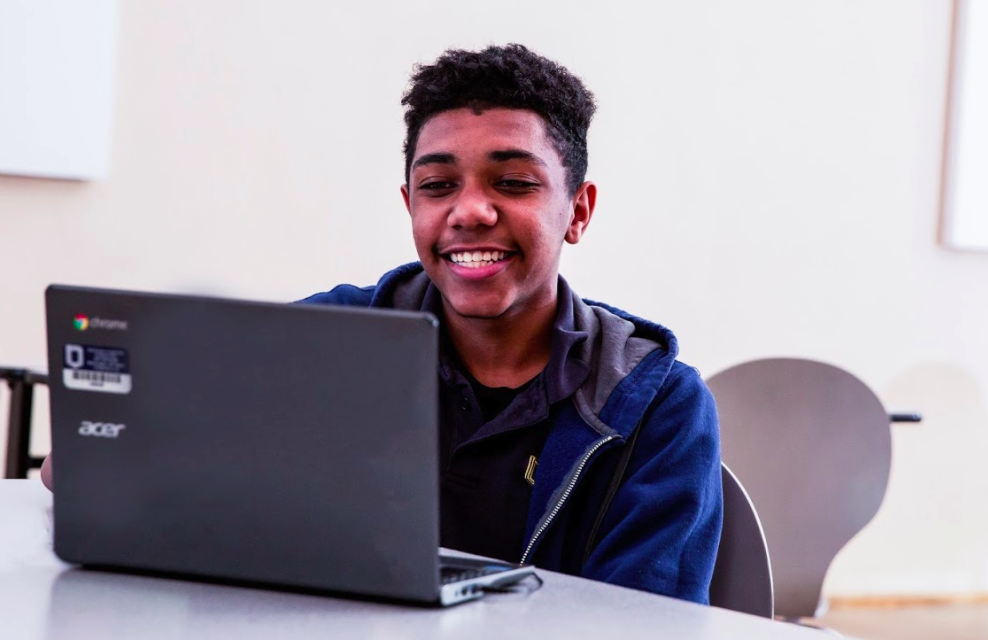 A student sitting at a table, looking at his computer screen, and smiling. 