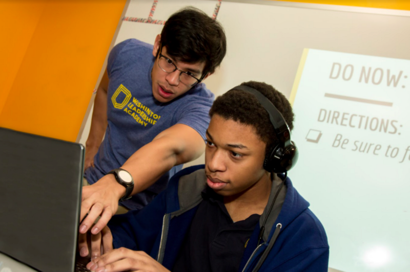 A student sitting and working on their computer. The teacher is standing next to him, pointing at the computer screen.