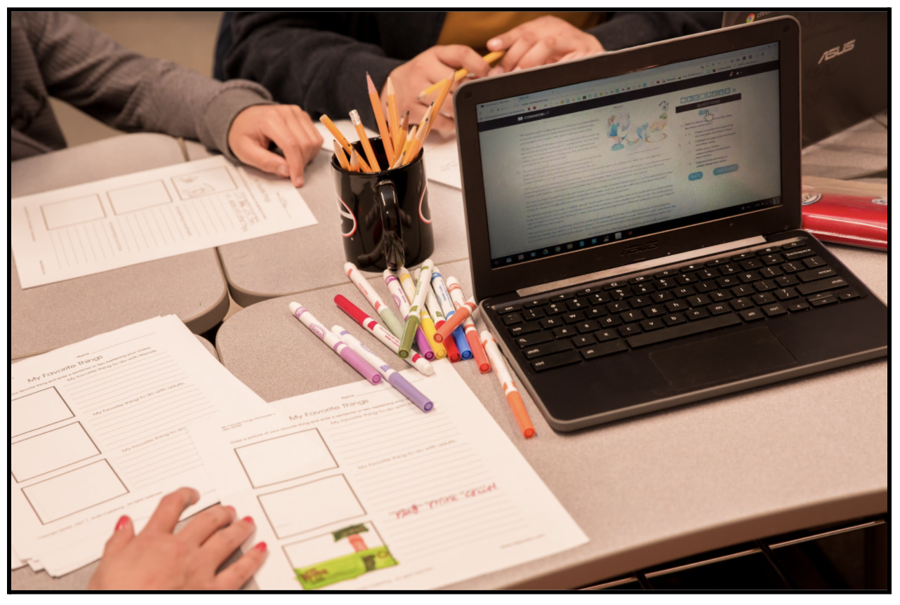 A table with printed graphic organizers, a pencil cup, markers, and a student's laptop. 