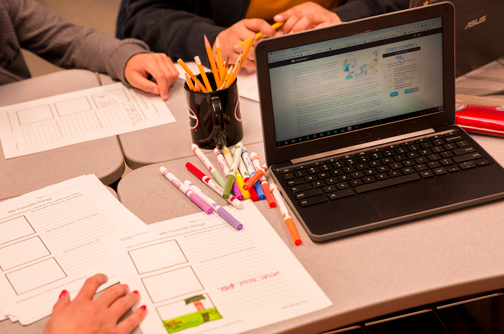A table with a computer open to a CommonLit lesson, printed graphic organizers, a pencil cup, and colorful markers.