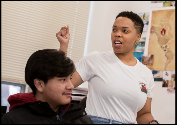 A student sitting next to a teacher who is standing up and gesturing. 