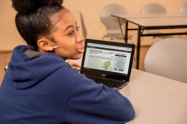 A student sitting at a table with a CommonLit lesson on her computer. She is looking off-camera. 