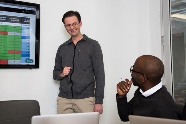 A man, standing, is looking at another man, sitting. The man sitting is looking at a data chart on a TV screen.