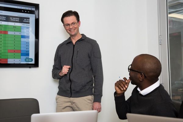 Two men look at each other in front of a TV screen with a data report. 
